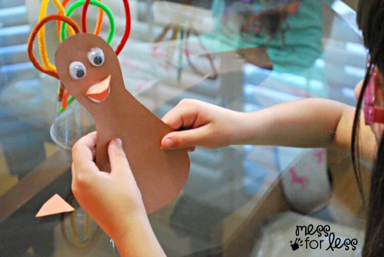 A child's hands holding brown construction paper in the shape of a turkey body with google eyes, orange construction paper beak, and different color pipe cleaners for the feathers.