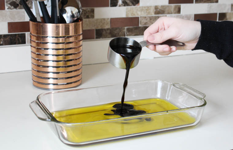A hand pouring balsamic vinegar into a clear baking dish that has olive oil in it.