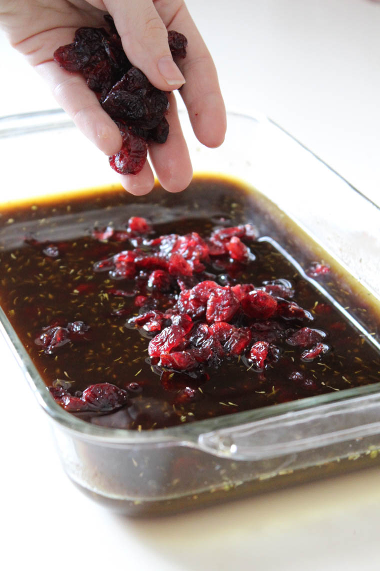 A hand dropping dried cranberries into a baking dish that has the marinade ingredients for cranberry balsamic chicken in it.