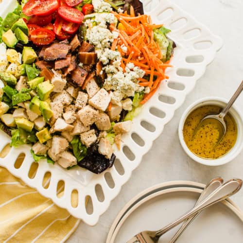 cobb salad on a white plate with yellow napkin and dressing on the side