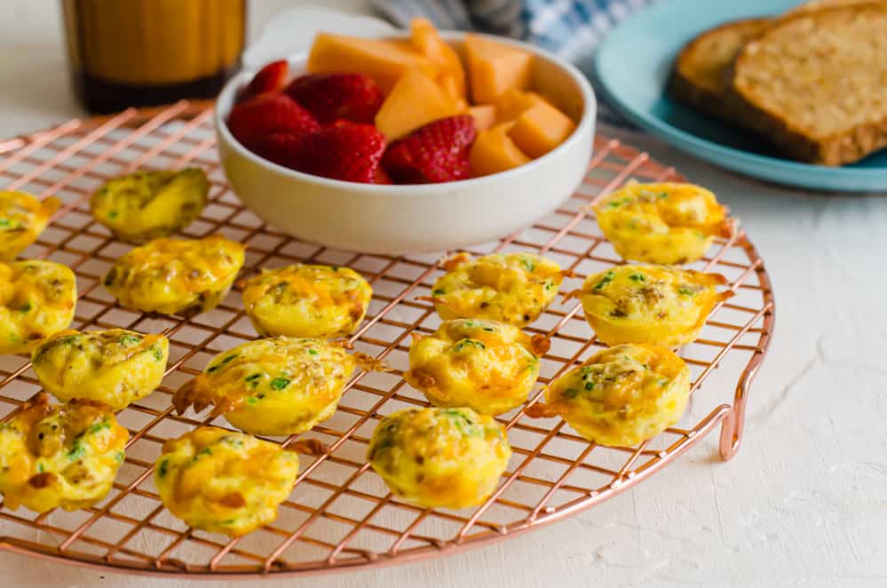 Cheddar chive egg bites on a baking rack with a bowl of fruit nearby.