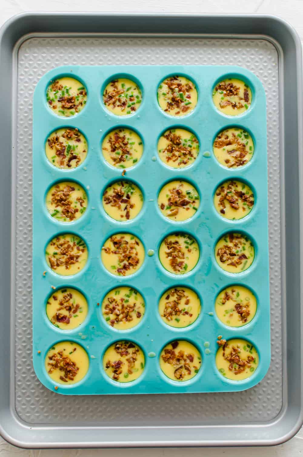 Beaten eggs with chopped chives and bacon pieces in a silicone mini muffin tin.