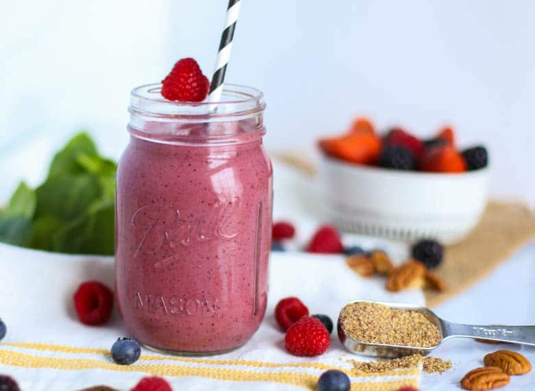 A very berry smoothie in a mason jar with a raspberry on the rim, a black and white striped straw in it, a measuring spoon of flaxseed next to it, and pecans, blueberries, blackberries, and raspberries scattered around.