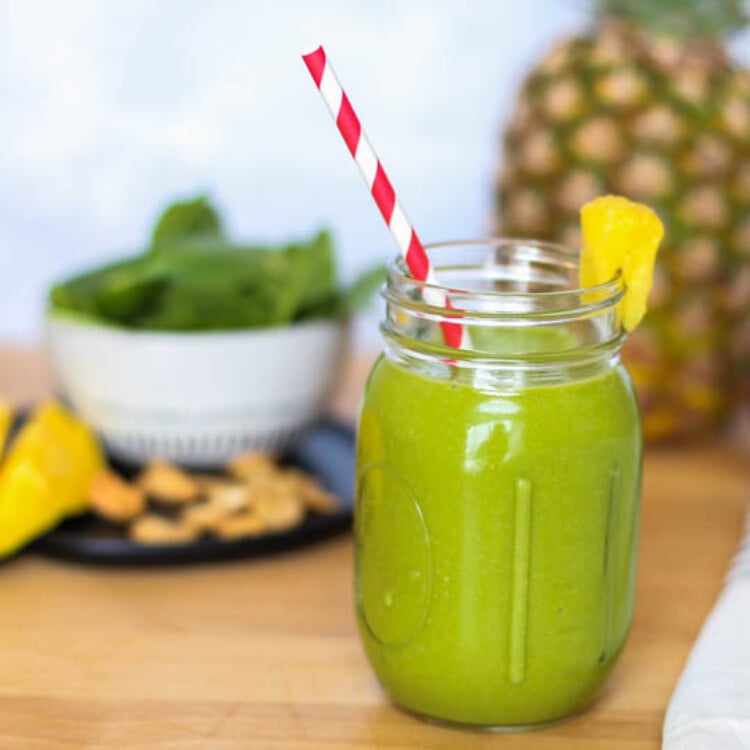 A green tropical smoothie in a mason jar with a bowl of spinach and a whole pineapple in the background.