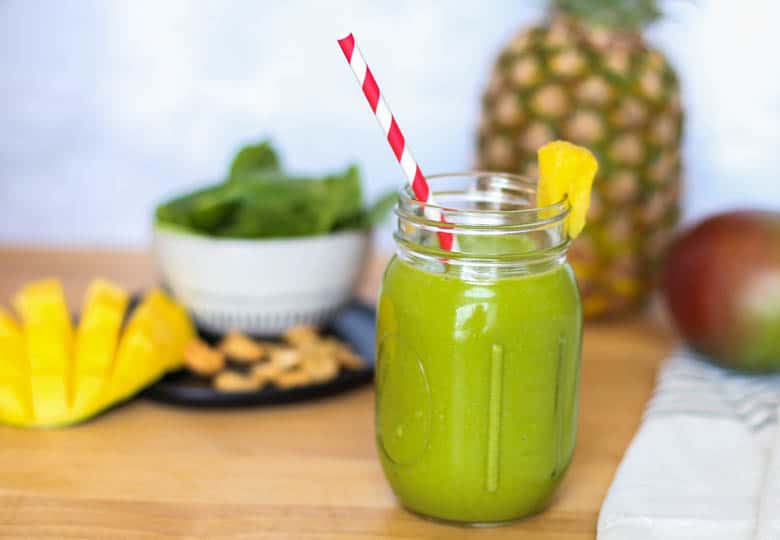 Tropical smoothie in a mason jar wit a pineapple chunk on the rim, a red and whtie striped straw in it, and spinach, cashews, mango, and pineapple in the background.