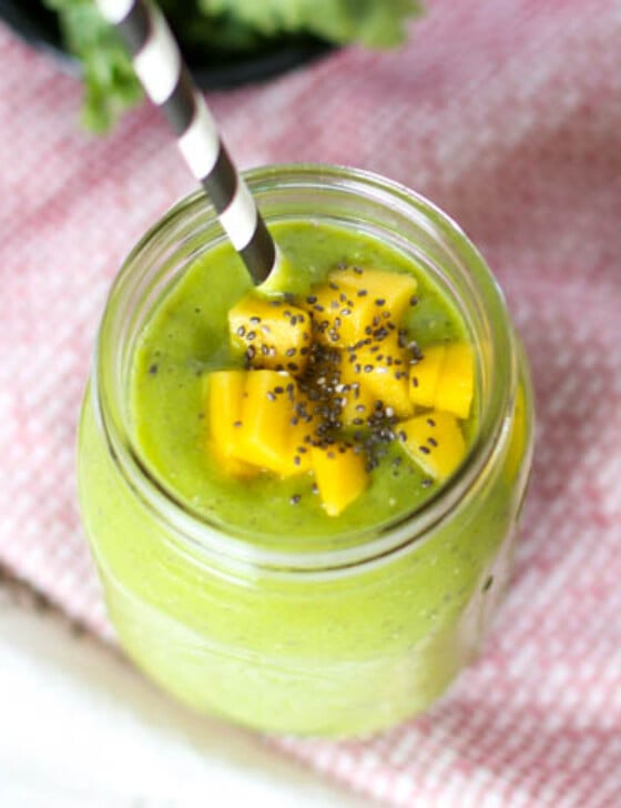A green smoothie in a mason jar with a measuring spoon of chia seeds and a sliced mango sitting next to it.