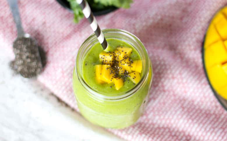 A green smoothie in a mason jar with a measuring spoon of chia seeds and a sliced mango sitting next to it.