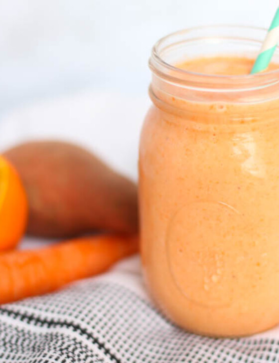 Orangesicle smoothie in a mason jar with a straw in it and a sweet potato, carrot, and half an orange sitting next to it.