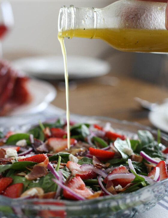 Lemon honey dressing being poured onto a spinach salad with sliced strawberries and red onions on top.