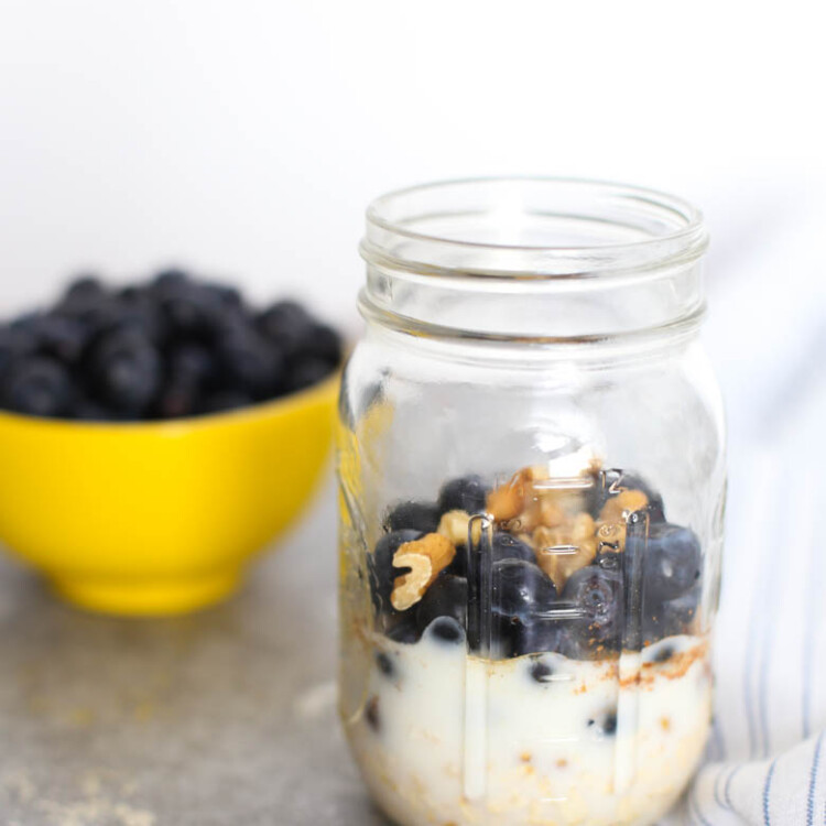 Blueberry muffin overnight oats ingredients layered in a mason jar with a bowl of blueberries in the background.