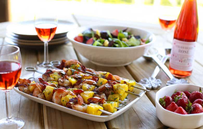 Bacon-wrapped shrimp kabobs on a platter and a bowl of strawberries pm a picnic table.