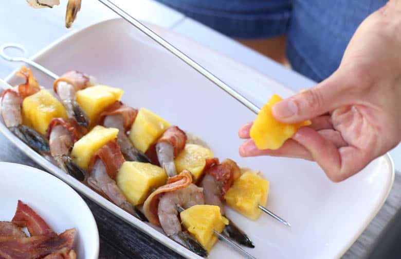 Someone skewering pineapple with two full bacon-wrapped shrimp kabobs.