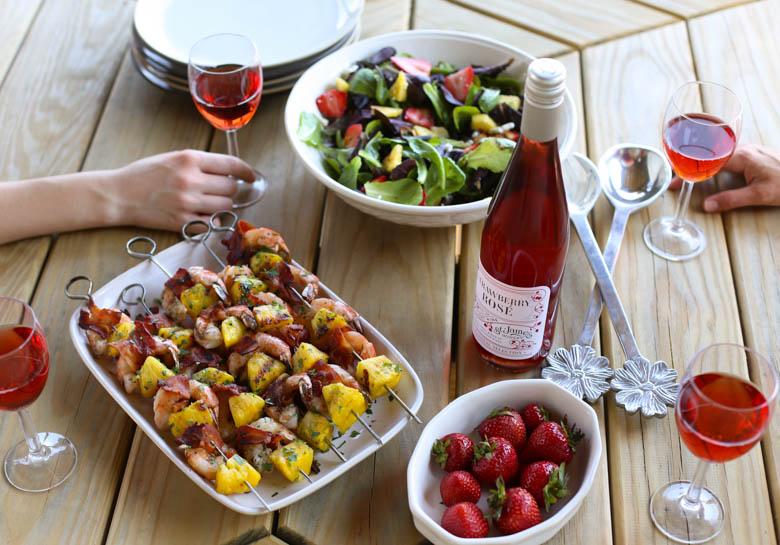 bacon-wrapped shrimp kabobs, a bottle of St. James Winery Strawberry Rose, strawberries, and green salad on a picnic table