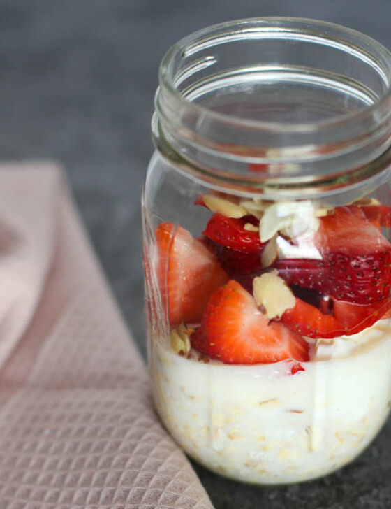 Strawberry shortcake overnight oats ingredients layered in a mason jar on a countertop with whole strawberries in the background.