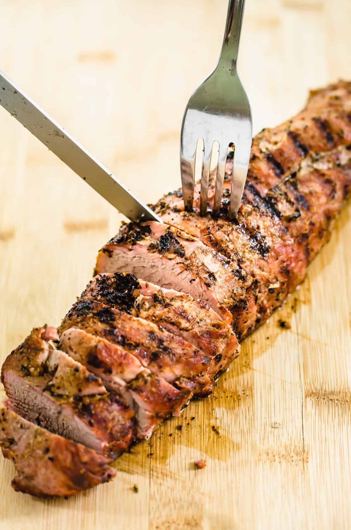 Roasted pork tenderloin with seasoning rub being sliced on a wooden cutting board.