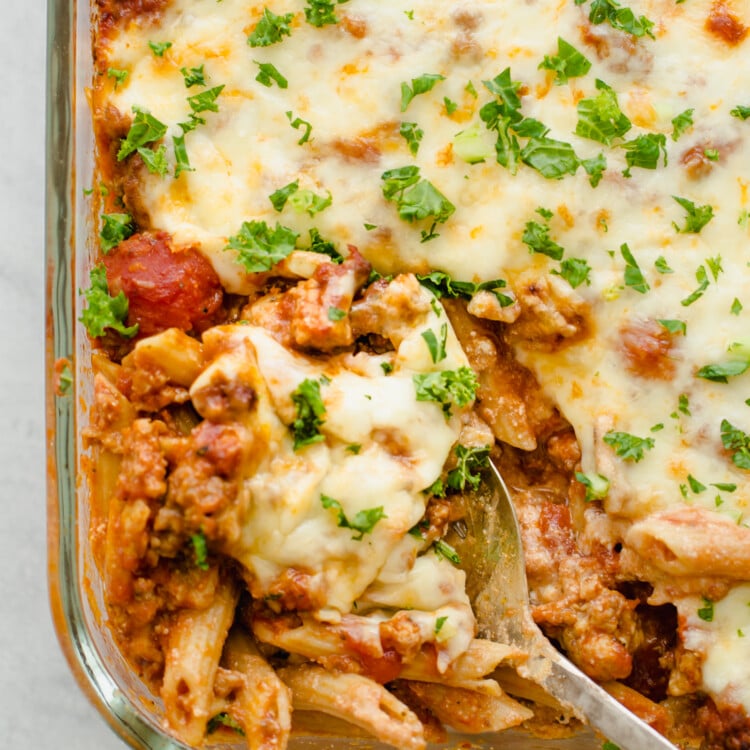 Baked penne pasta being scooped out of a glass baking dish.