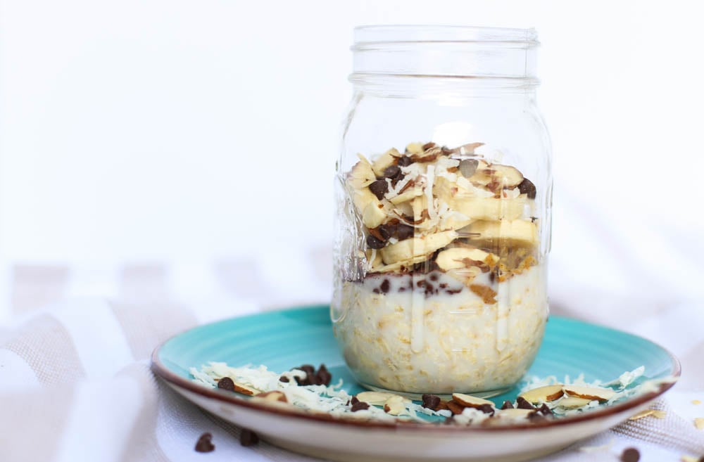 Almond Joy overnight oats in a mason jar sitting on an aqua plate with mini chocolate chips, shredded coconut, and sliced almonds sprinkled around the plate and table.