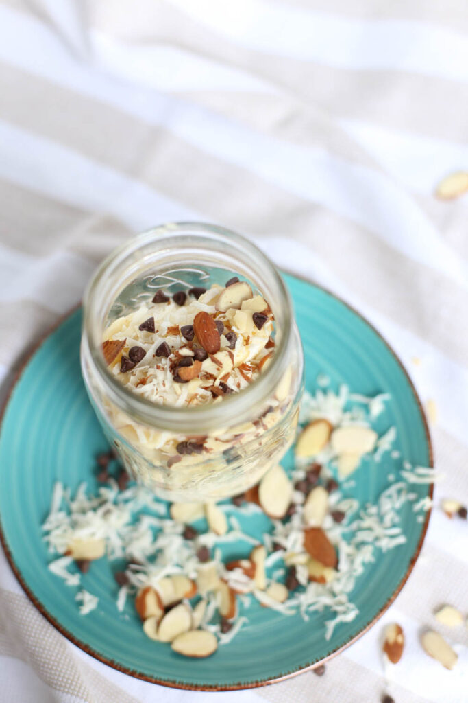 Almond Joy Overnight Oats in a mason jar on top of turquoise plate. Shredded coconut, sliced almonds, and chocolate chips are scattered around.