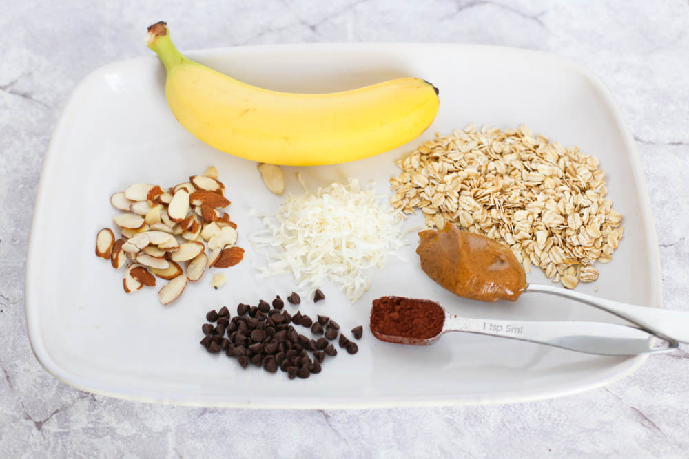 Almond Joy Overnight Oats ingredients on a white plate. Includes bananas, oats, almond butter, cocoa, chocolate chips, shredded coconut, and sliced almonds.