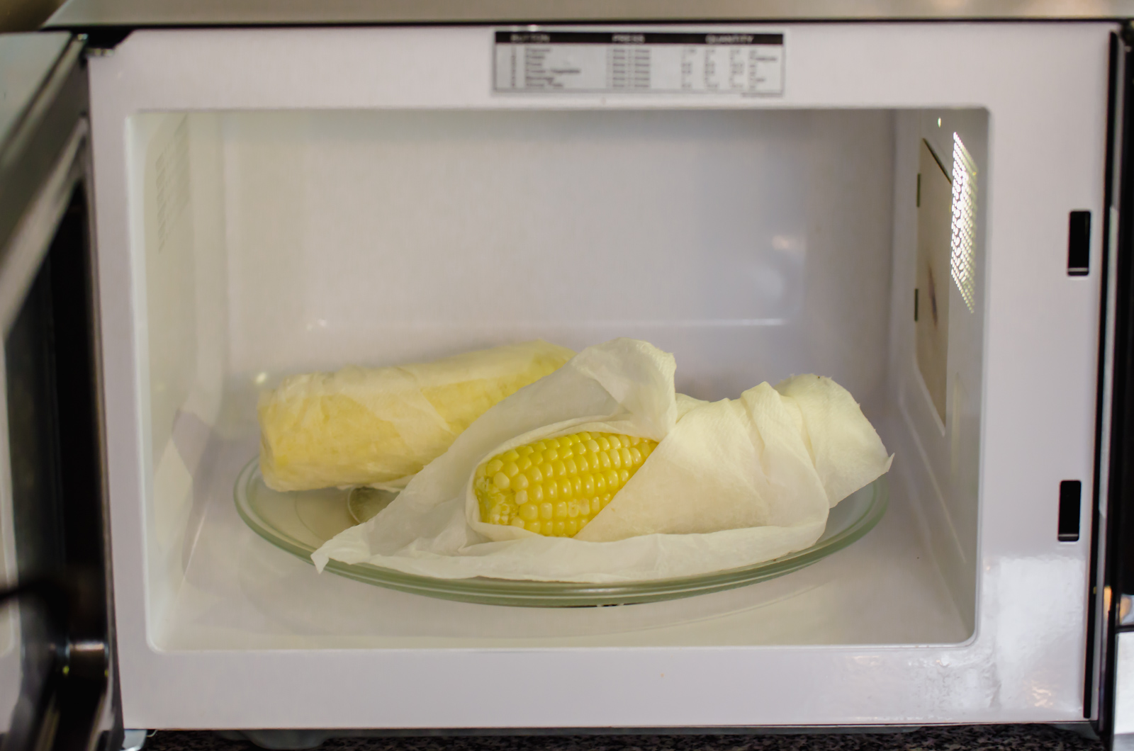 Two ears of corn on the cob wrapped in damp paper towels sitting in a microwave.