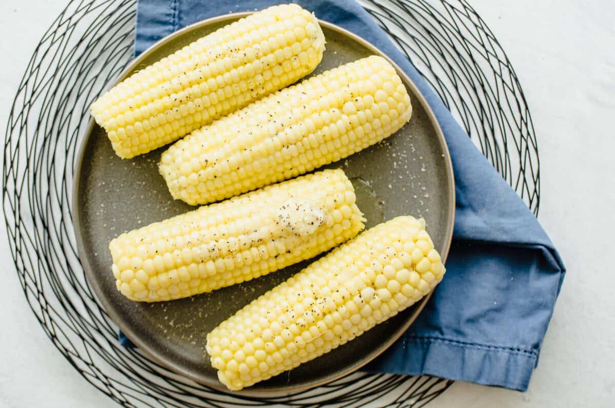 Four ears of corn on the cob on a plate with butter, salt, and pepper on top.