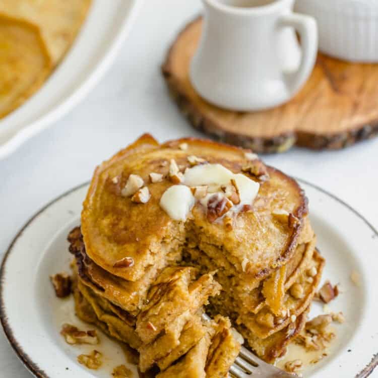 Whole wheat pumpkin pancakes stacked on a plate with pecans, butter, and syrup on top.