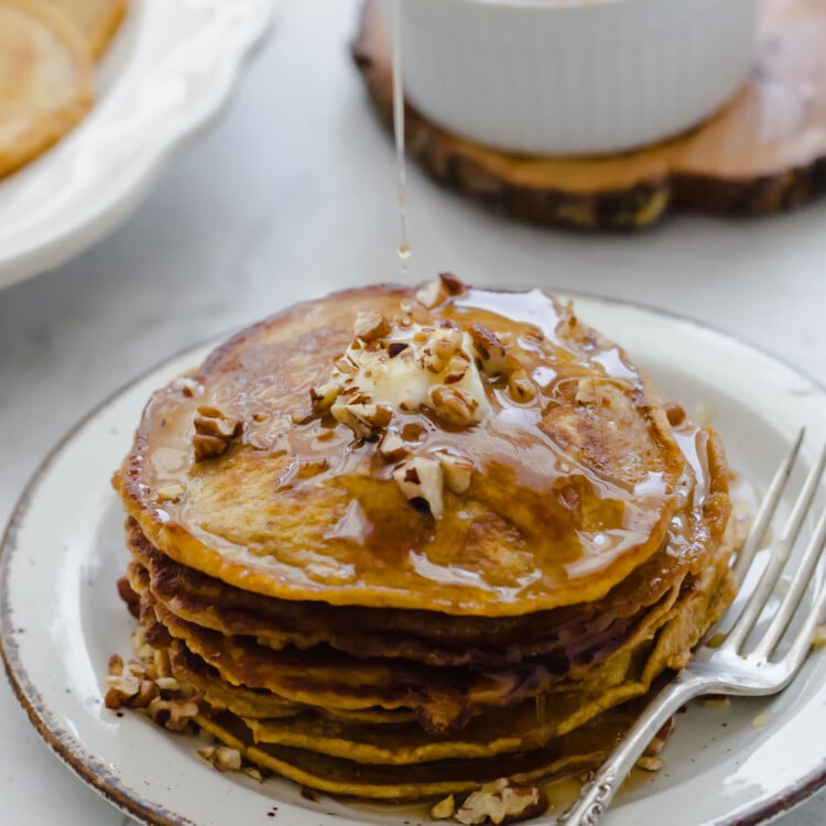 Whole wheat pumpkin pancakes stacked on a plate with syrup being poured on top.