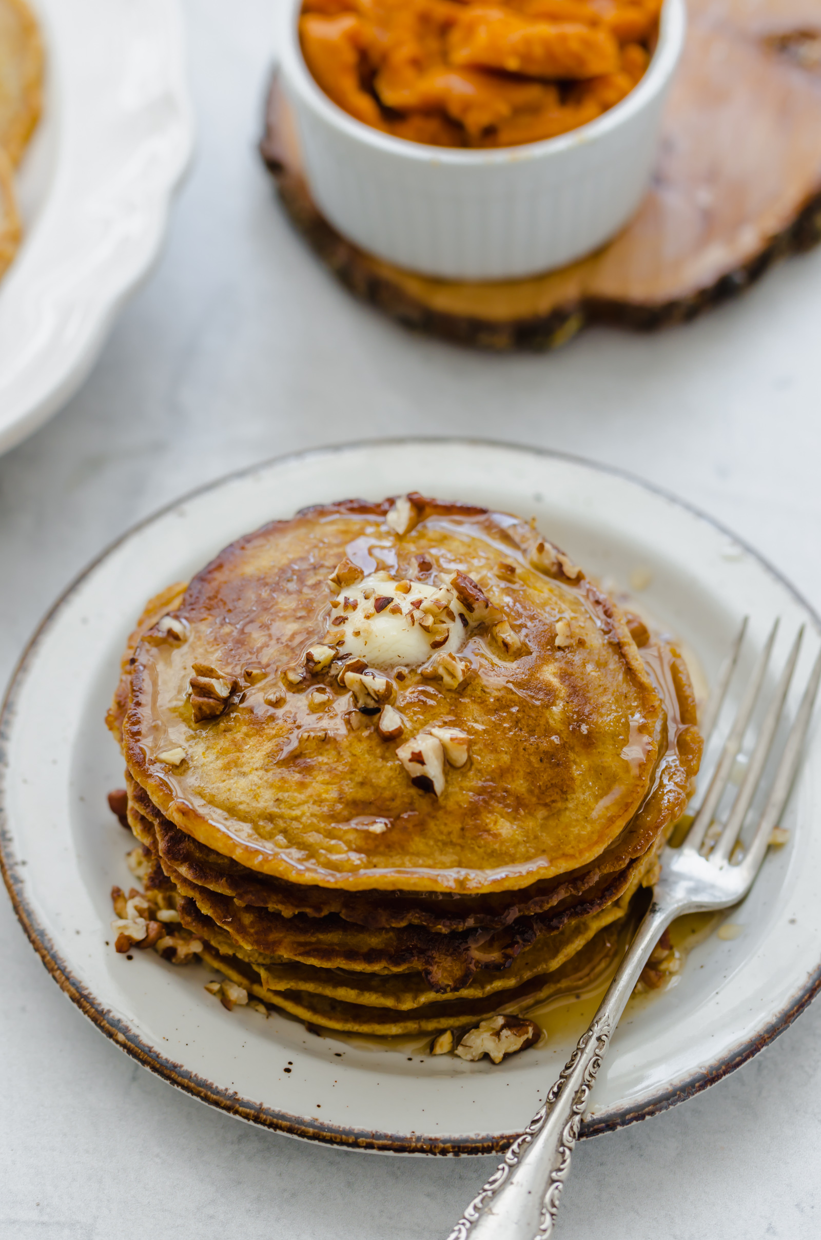 Whole wheat pumpkin pancakes stacked on a plate with butter, syrup and chopped pecans on top.