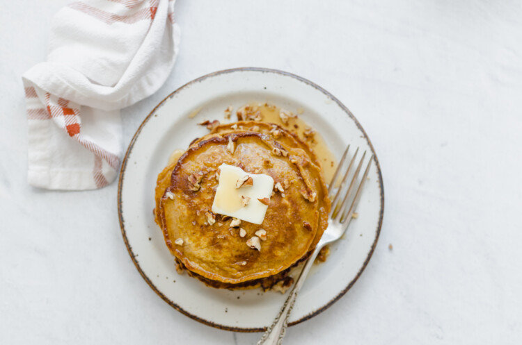 Pumpkin pancakes stacked on a plate with syrup, a pat of butter, and pecan pieces on top.