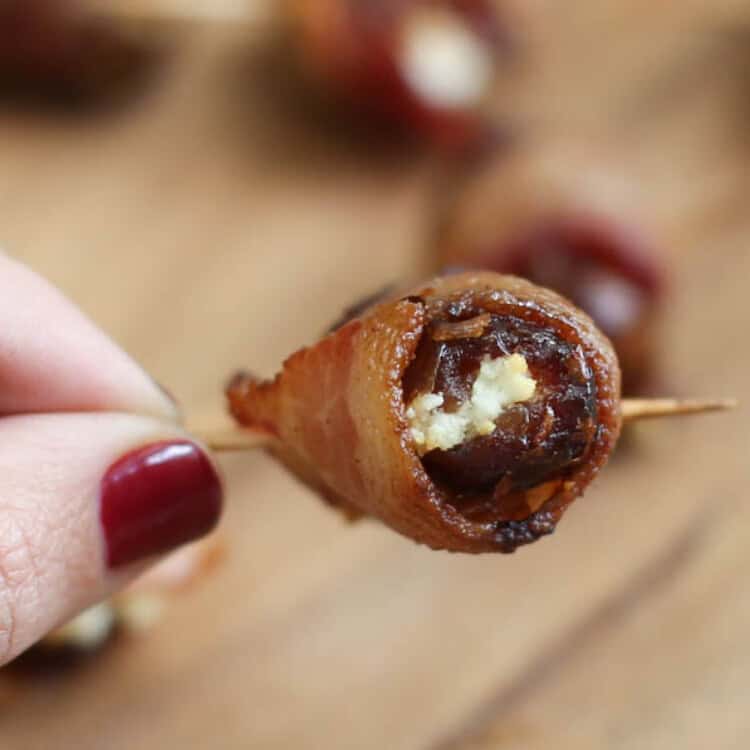 A hand holding the toothpick of a fully-cooked Bacon-Wrapped Date with Goat Cheese.