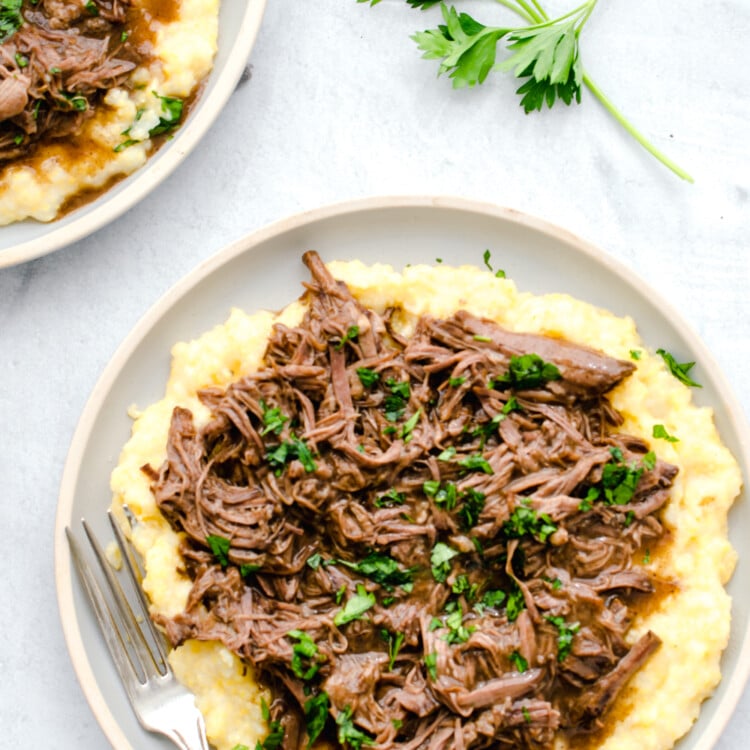 Balsamic shredded beef on top of polenta on a white plate with fresh chopped parsley on top.