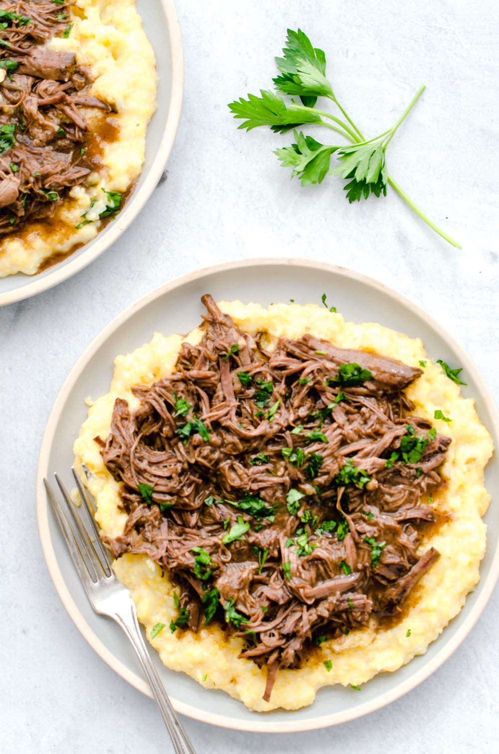 Balsamic shredded beef on top of polenta on a white plate with fresh chopped parsley on top.