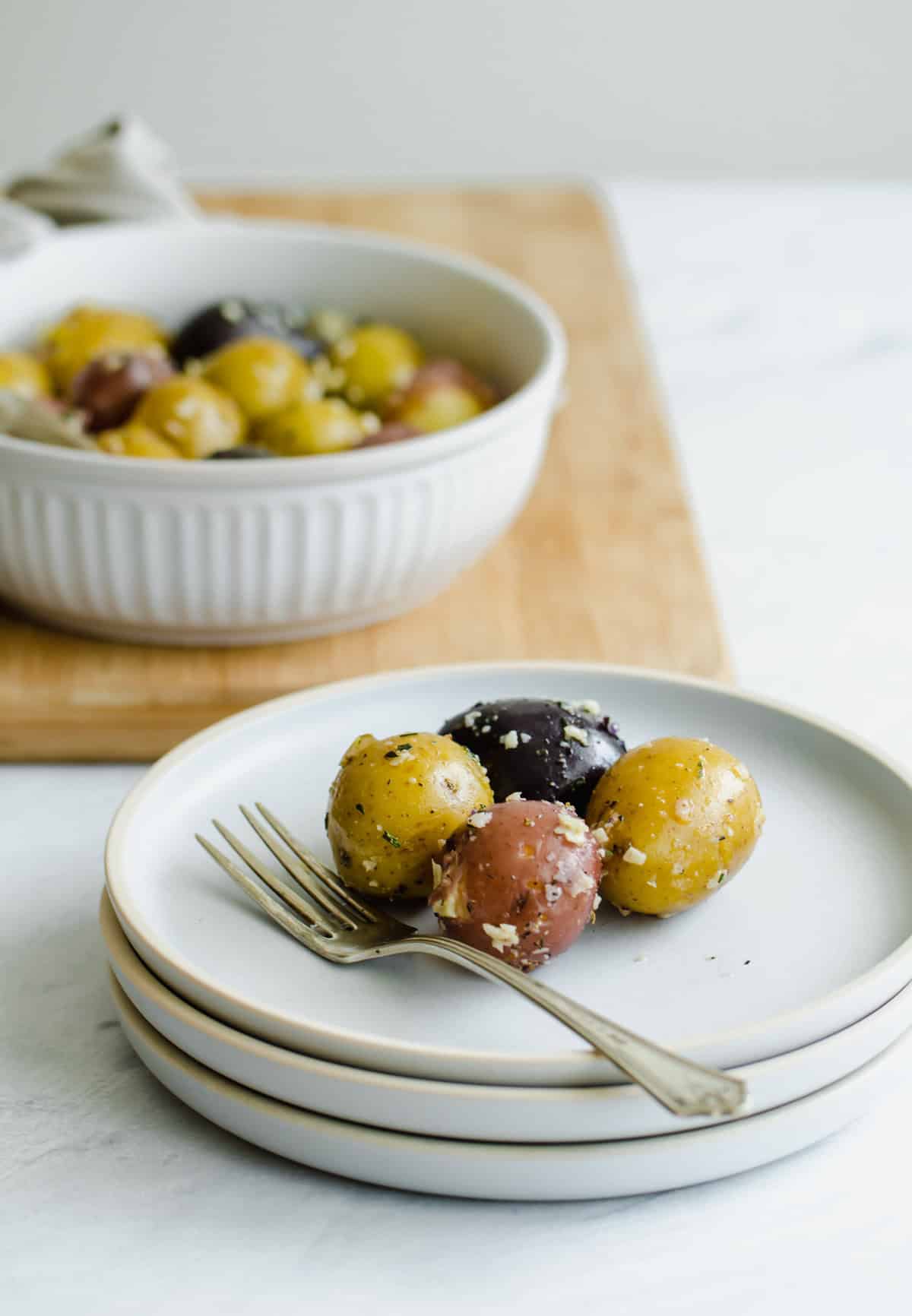 Four boiled baby potatoes on a stack of white plates with a serving dish of boiled baby potatoes in the background.