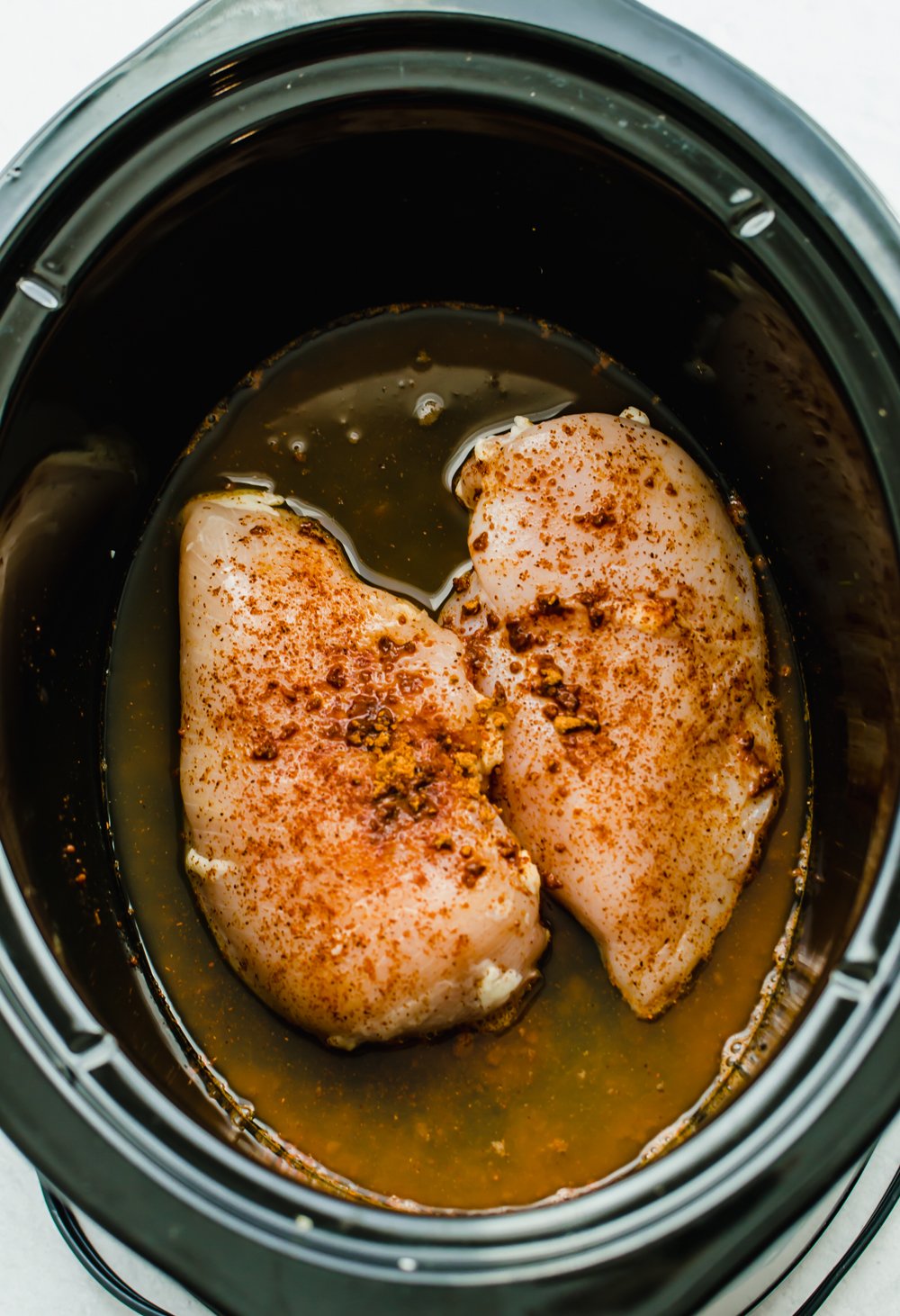 Raw chicken breasts with Mexican seasonings in a slow cooker with broth around it.