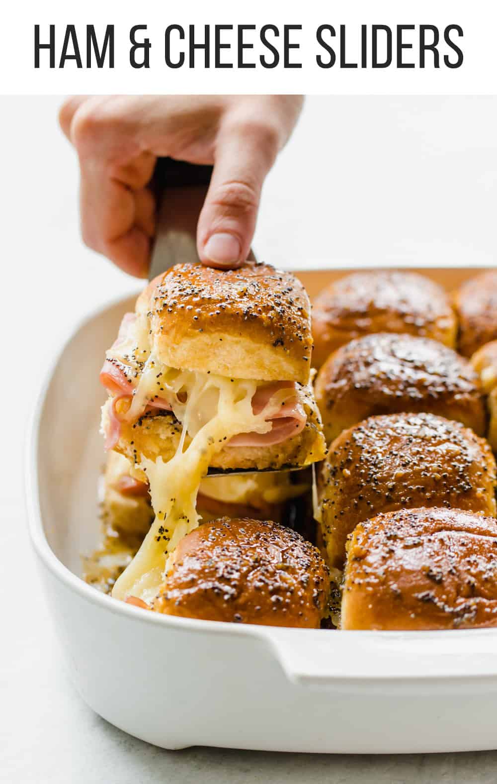 Hand lifting a ham & cheese slider from a white dish with a spatula.