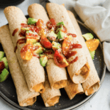 Creamy chicken taquitos stacked on a plate with avocado and tomato chunks on top.