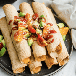 Creamy chicken taquitos stacked on a plate with avocado and tomato chunks on top.