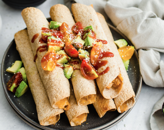 Creamy chicken taquitos stacked on a plate with avocado and tomato chunks on top.