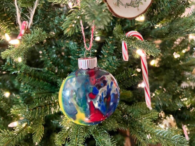 A clear ornament with paint swirled inside hung on a Christmas tree next to a candy cane.