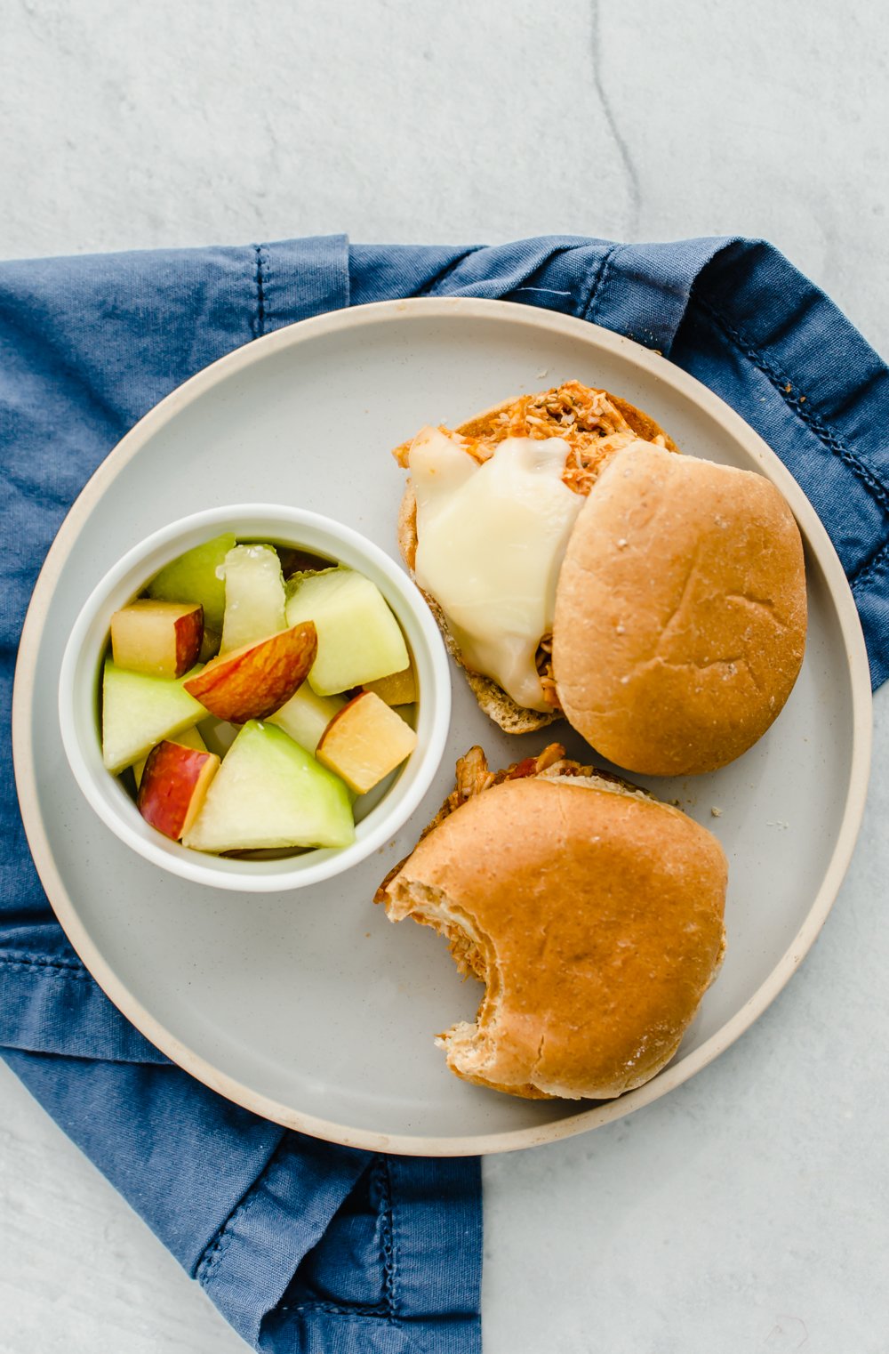Two chicken Parmesan sliders on a white plate with a bowl of cut up fruit.