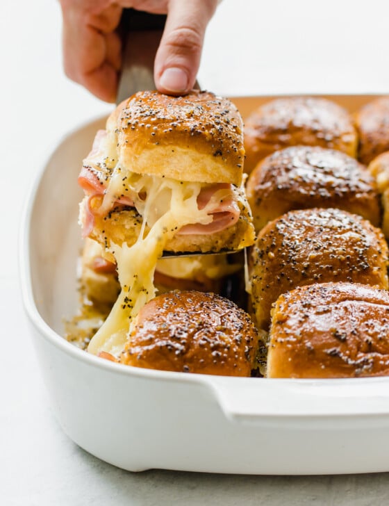 A hand lifting a ham and cheese slider from a baking dish with a spatula.