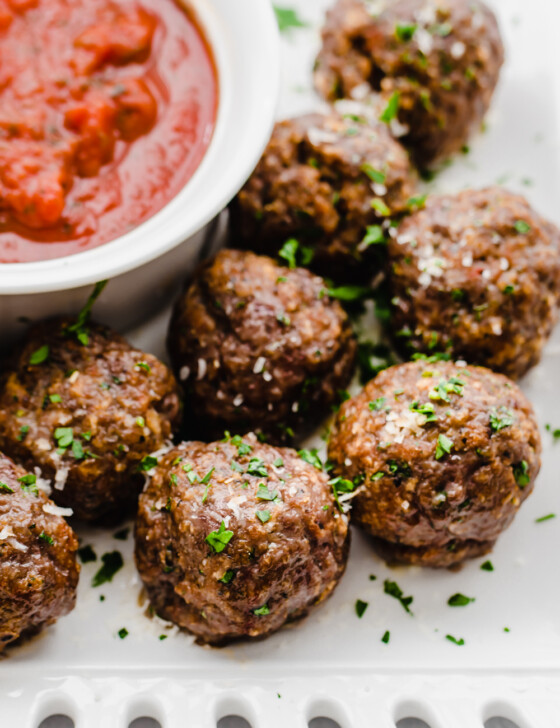 Baked Italian meatballs on a white platter with a small bowl of marinara sauce.