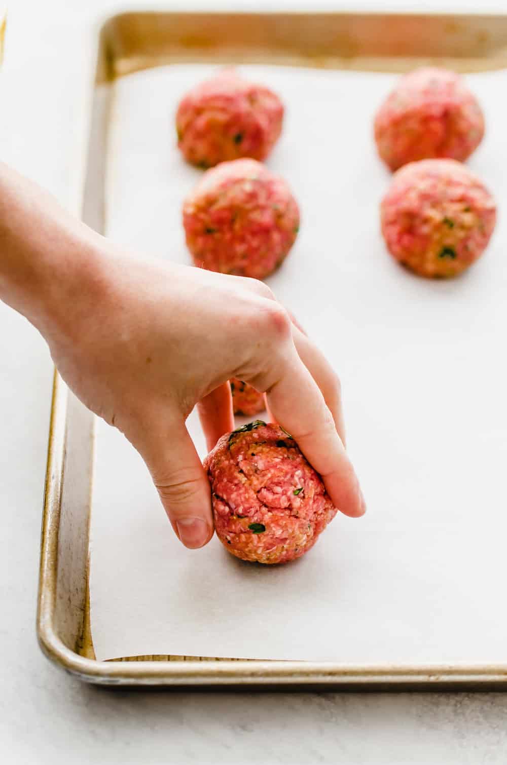 A hand placing a raw meatball on a parchment-lined baking sheet.