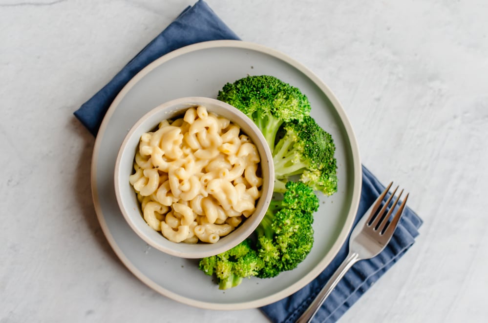 Mac n cheese in a bowl sitting on a gray plate with steamed broccoli on the side.