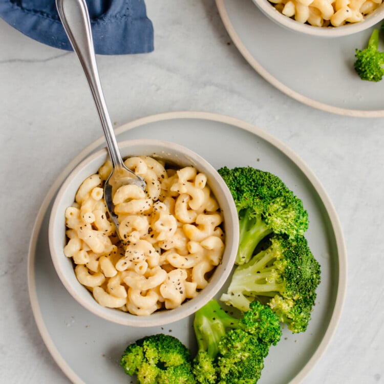 Mac and cheese in bowls on white plates with steamed broccoli on the side.