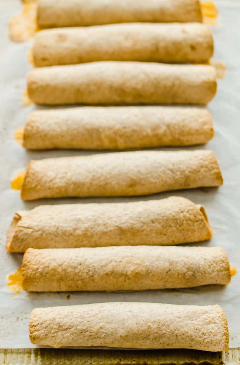 slow Cooker Chicken and Cheese Taquitos are rolled up and lined up on parchment paper.