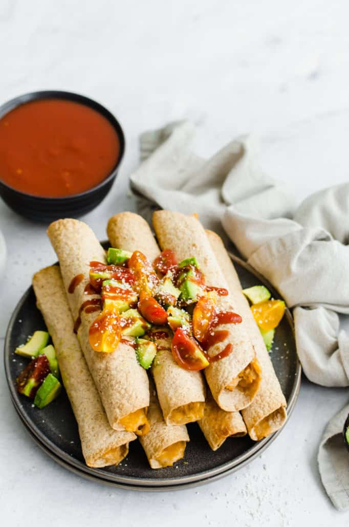 Taquitos cu pui și brânză îngrămădiți pe un platou de servire cu salsa de avocado și lime deasupra și salsa alături.