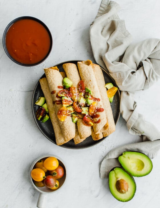 Chicken and Cheese Taquitos piled on a serving platter with avocado lime salsa on top. Other toppings are nearby, like avocado, tomatoes, and salsa.