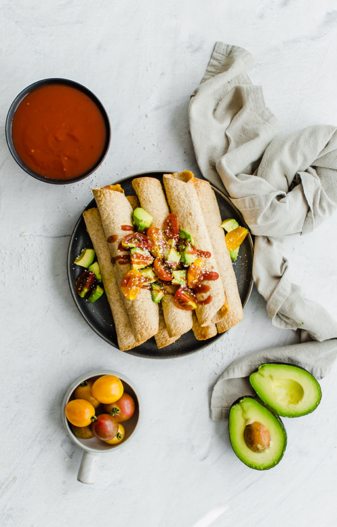 Chicken and Cheese Taquitos piled on a serving platter with avocado lime salsa on top. Other toppings are nearby, like avocado, tomatoes, and salsa.