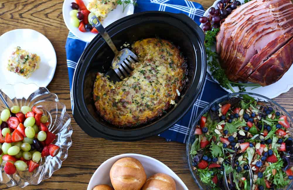 Slow cooker with hash brown casserole, a ham, salad, fruit salad, and plates on a wooden table